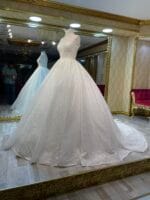 Aygök Gelinlik Prenses Simli Kumaş kabarıkk - VznModa White ballgown on a mannequin in a luxurious bridal shop with gold-trimmed mirrors and patterned walls, a second dress reflected in the mirror behind, and a man taking a photo in the background