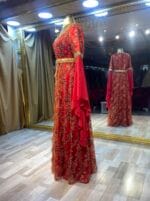 Vina kırmızı gold payet bindallı kına abiye elbiseee - VznModa Red floor-length gown with gold embroidery on a mannequin in a boutique window display, featuring a gold belt and draped red cape sleeve.
