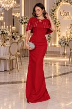 Woman in a red evening gown with cape-like shoulders posing in a chandelier-lit ballroom, holding a bejeweled silver clutch.
