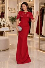 Woman in a red evening gown with a beaded cape standing in a luxury bridal boutique, holding a silver clutch.