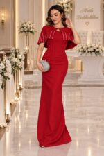 Elegant woman in a red gown posing in a wedding venue adorned with white flowers and soft lighting, holding a jeweled clutch.