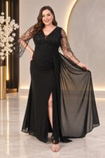 Hercai siyah büyük beden dantel şifonn abiye elbise - VznModa Woman in a black evening gown with beaded sheer sleeves, posing with one hand on hip in a elegant lobby.